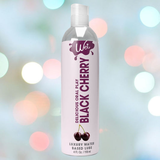 A transparent bottle of Wet Delicious Oral Play Black Cherry water-based flavored lubricant prominently displays the name "Delicious Oral Play Black Cherry" against a backdrop adorned with black cherry designs and soft, colorful bokeh lights. 1080