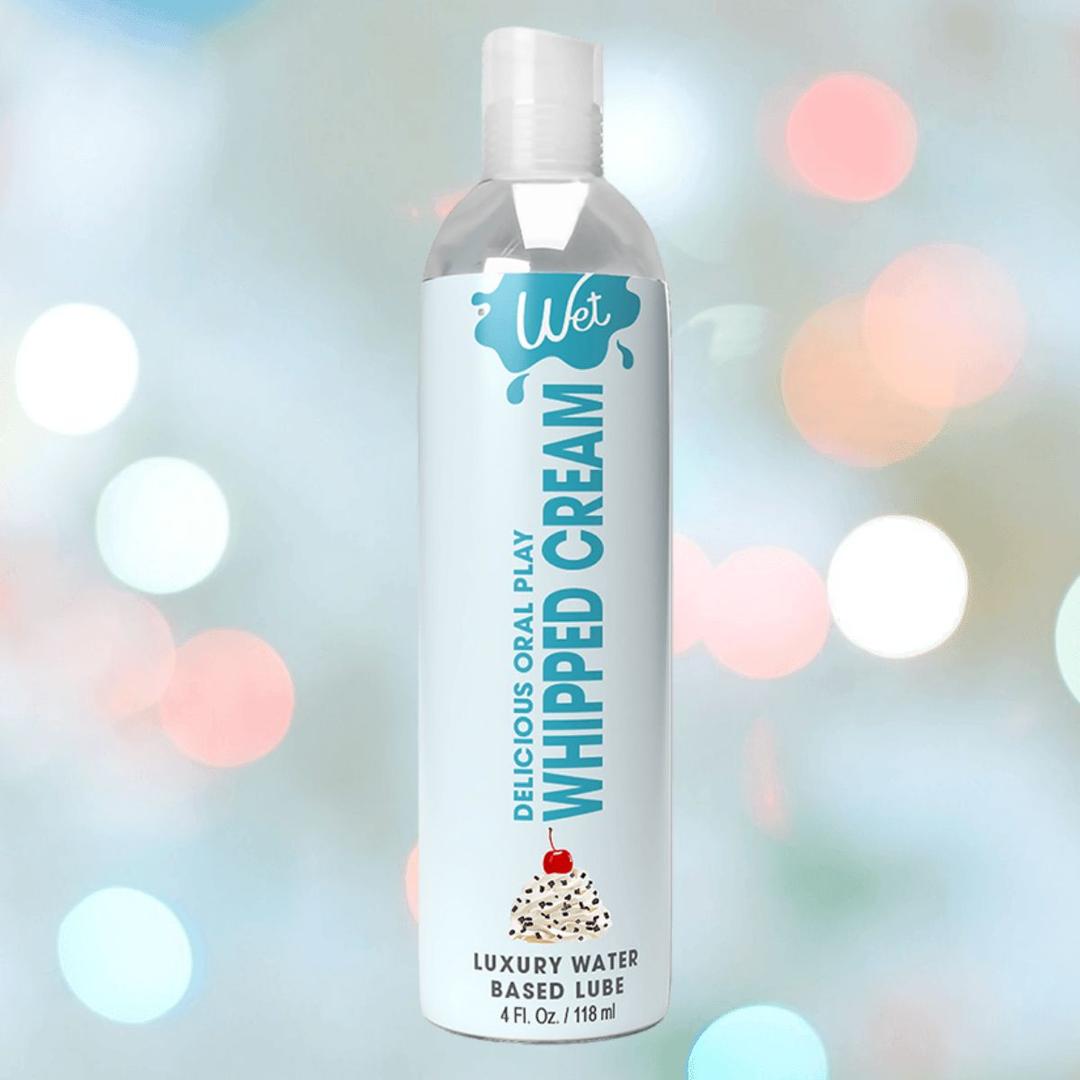 A 3oz bottle of Wet Desserts Whipped Cream Flavored Lubricant by Wet sits against a colorful, blurred background with sprinkles and light bokeh effects.