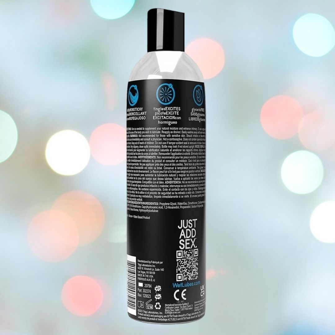 A black bottle with a black cap and white-blue text stands against a colorful blur. The label reads "JUST ADD SEX" and features Wet Extra Sensations Tingling Water/Silicone Blend Based Lubricant by Wet.