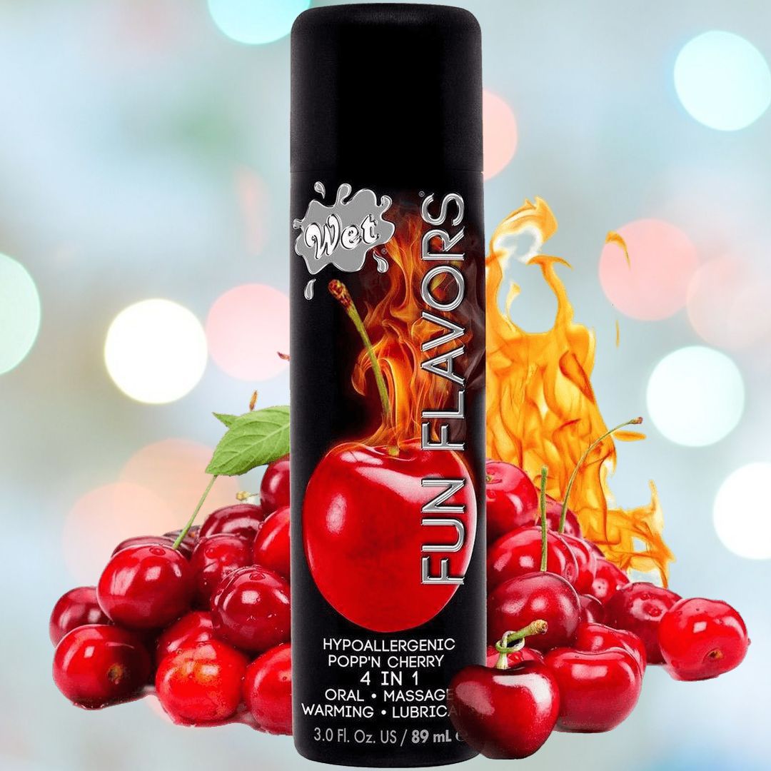 The image features a black bottle with a vibrant label that reads "Wet Fun Flavors" and "Hypoallergenic Popp'n Cherry, 4-in-1, Oral, Massage, Warming, Lubricant." In the background, you can see cherries and an explosion of flames against a soft-focus backdrop. This cherry-flavored warming lubricant by Wet is perfect for any mood.