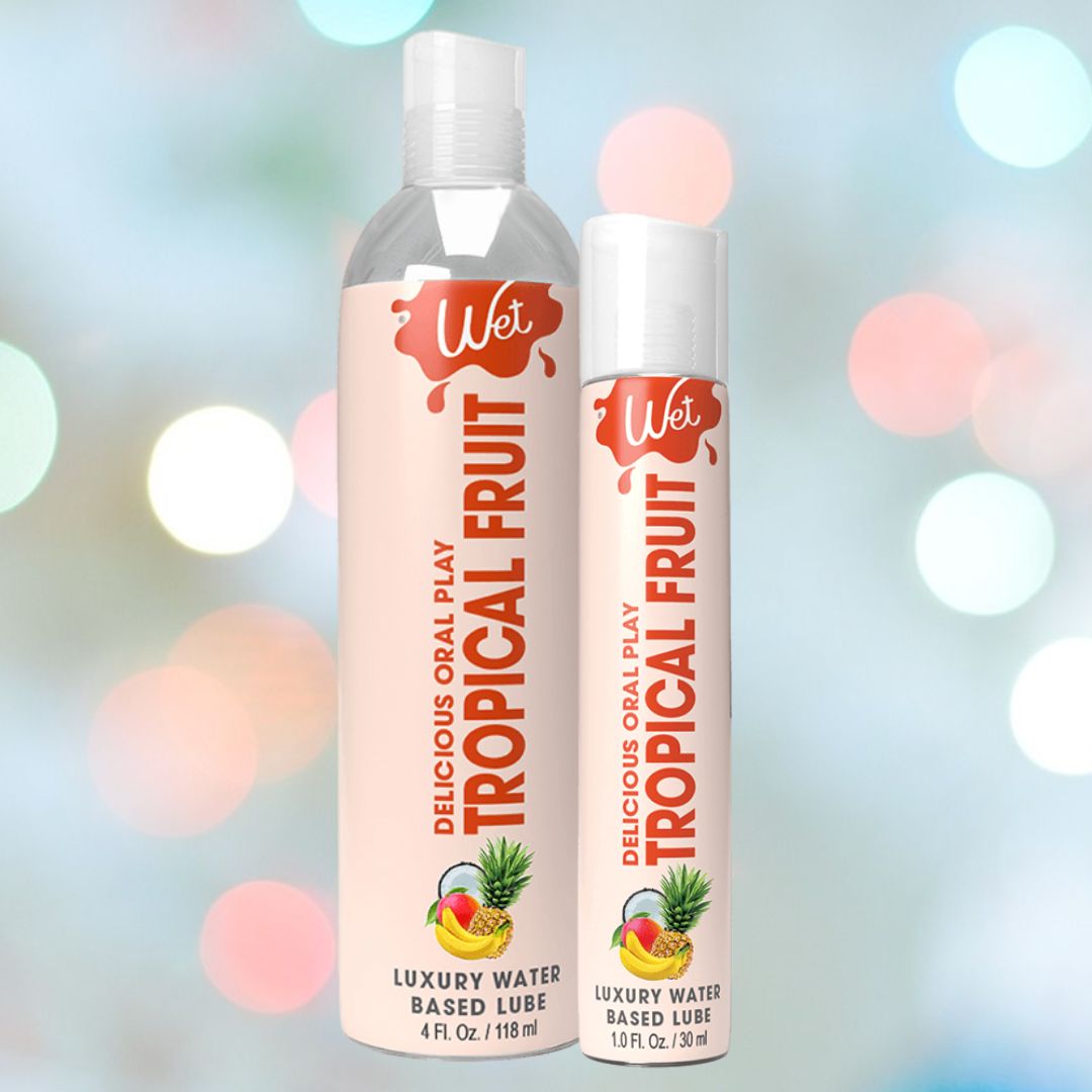 Displayed against a blurred background with circular light spots are two bottles of Wet "Tropical Explosion" flavored lubricant by Wet: one large, one small. Each bottle is adorned with a vibrant fruit graphic featuring pineapple and mango, highlighting the water-based formula's tropical essence.