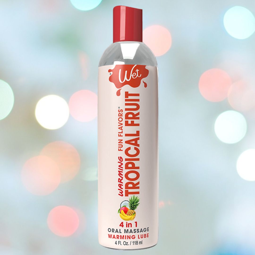 A bottle of Wet "Tropical Explosion" Warming Lubricant 🍍 is showcased against a blurred background filled with multicolored lights. The mostly white bottle, adorned with red text and a tropical fruit graphic, promises a water-soluble experience.