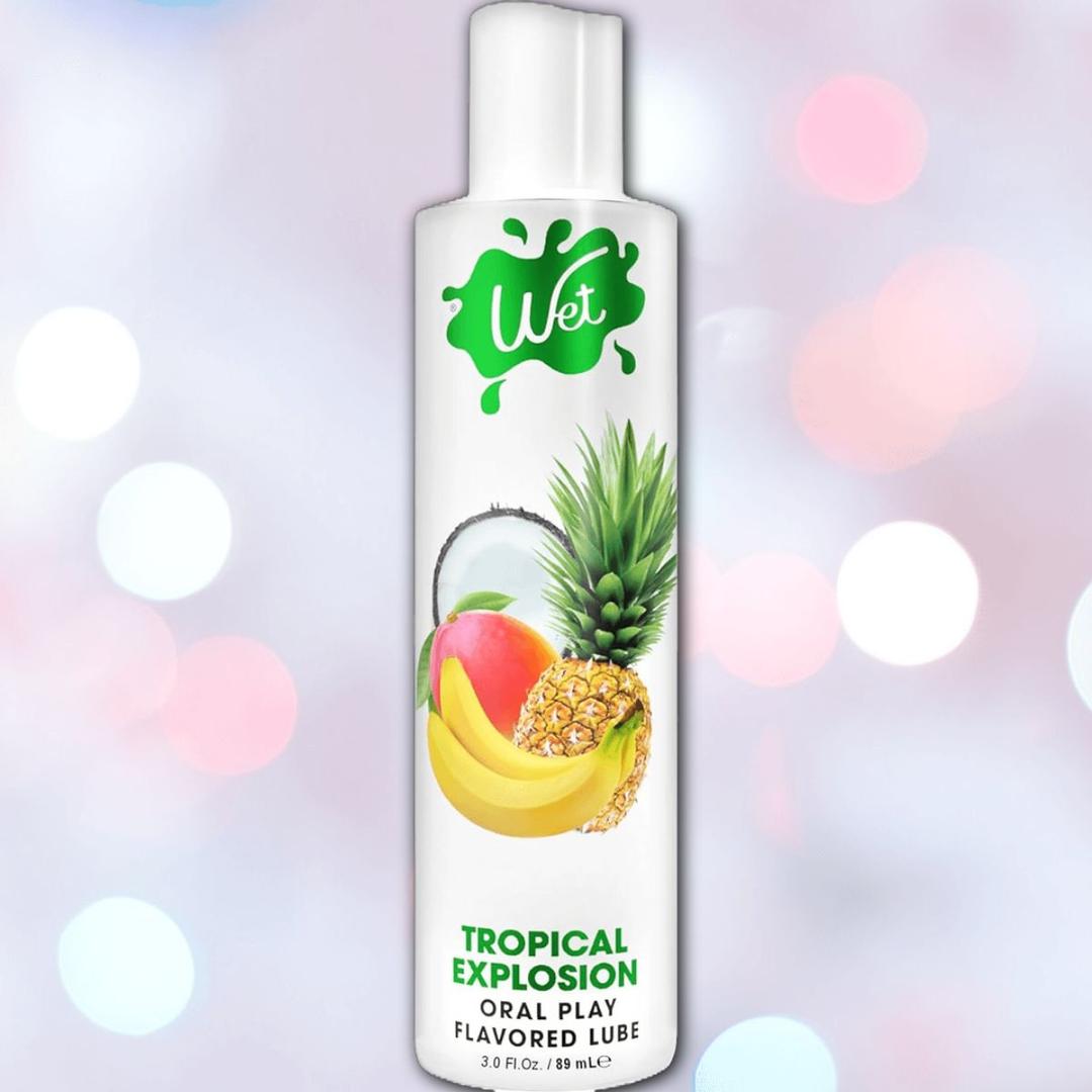 A bottle of Wet "Tropical Explosion" Warming Lubricant 🍍, with vibrant mango, banana, pineapple, and coconut images on the label set against a colorful bokeh background—perfect for a deliciously smooth tropical experience.