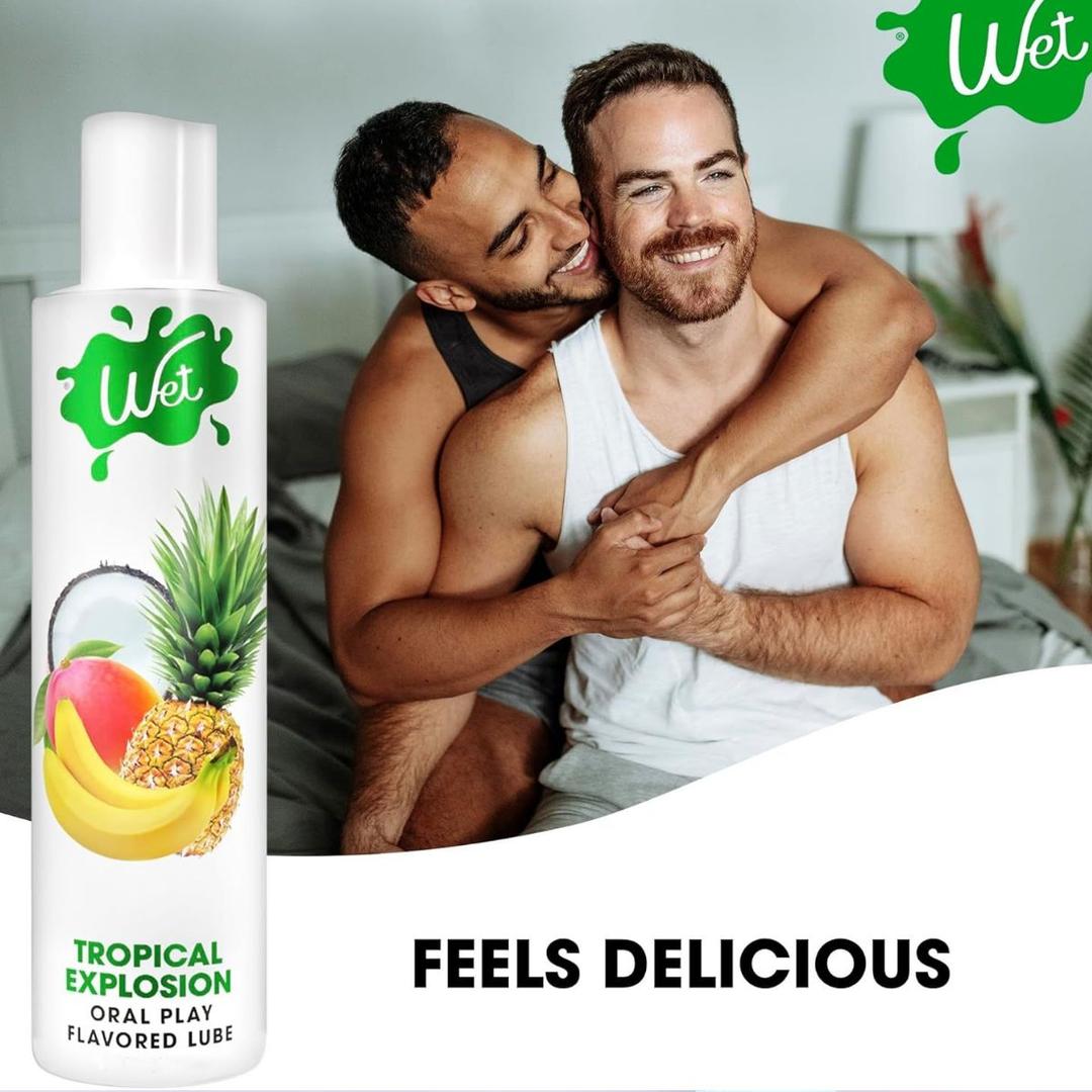Two men smile and embrace on a bed. In the foreground is a bottle of Wet "Tropical Explosion" Warming Lubricant 🍍 by Wet. The text below reads: “FEELS DELICIOUS.”.