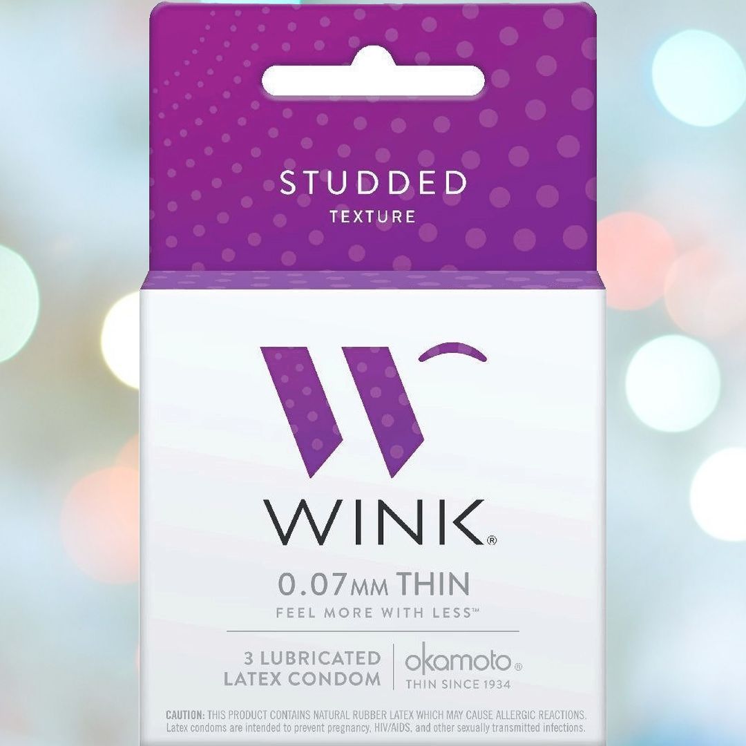 A box of "Okamoto Wink Studded Condoms" featuring raised rubber studs, 0.07mm thickness, and silicone lubricant. Each pack contains three condoms and the packaging boasts a purple and white minimalist design with colorful blurred lights in the background. The brand "Okamoto" is noted at the bottom.