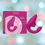 A pink Womanizer Blend Vibrant Air-Pulse Clit Stimulator by Womanizer is showcased against a backdrop of soft, colorful bokeh lights. The product packaging emphasizes features such as dual stimulation and adjustable intensity levels, along with G-spot vibrations for an enhanced experience.