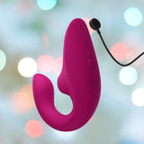 The Womanizer Blend Vibrant Air-Pulse Clit Stimulator in pink by Womanizer is showcased against a softly blurred background of colored lights, featuring buttons and a charging cable for enhanced G-spot vibrations and pleasure.