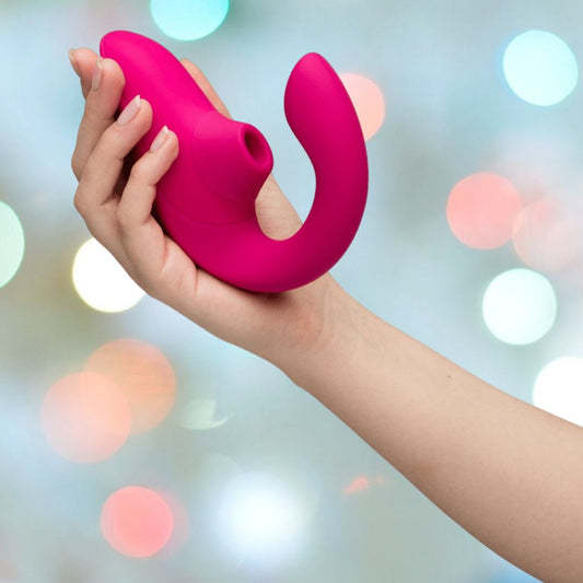 A hand holds the Womanizer Blend Vibrant Air-Pulse Clit Stimulator in pink against a softly blurred background with multicolored lights. 1080