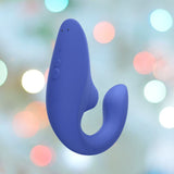 The Womanizer Blend Vibrant Air-Pulse Clit Stimulator in blue, crafted from curved silicone and featuring buttons for control, is intended for personal use. Displayed against a softly blurred backdrop with colorful bokeh lights, this product by Womanizer offers the promise of blended orgasms.