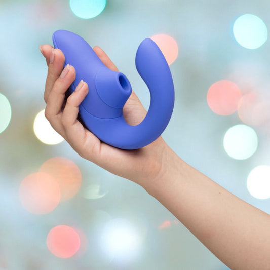 A hand holds a curved, blue, U-shaped Womanizer Blend Vibrant Air-Pulse Clit Stimulator against a softly blurred background with bokeh lights. The Womanizer product features a smooth surface with a small circular opening at one end. 1080