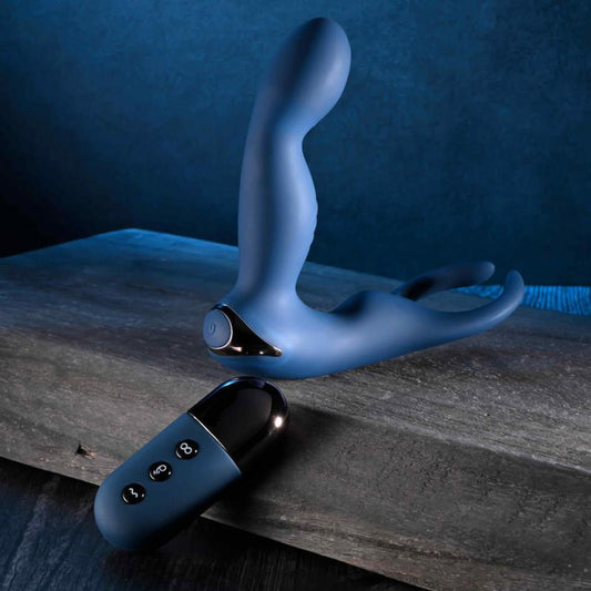 The Zero Tolerance By All Means Remote Control Prostate Vibrator, featuring a blue, flexible design with dual prongs, is positioned on a textured surface. In front lies a black remote control with three buttons that provide Turbo Mode excitement, all set against a dark blue background. 1080