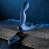 The Zero Tolerance By All Means Remote Control Prostate Vibrator, featuring a blue, flexible design with dual prongs, is positioned on a textured surface. In front lies a black remote control with three buttons that provide Turbo Mode excitement, all set against a dark blue background.