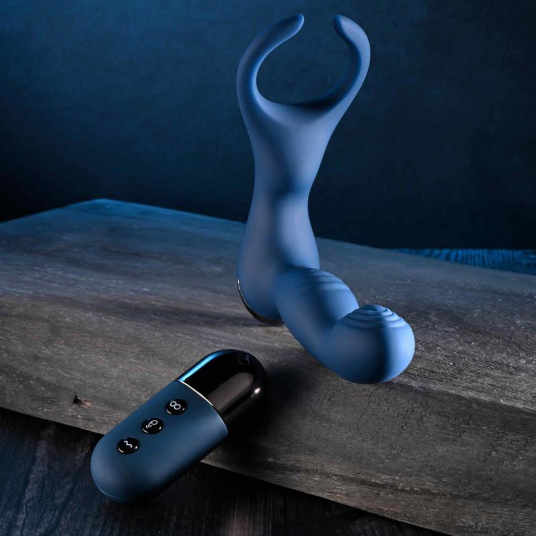 A sleek blue silicone wearable adult toy with a U-shaped opening and ribbed texture lies beside the Zero Tolerance By All Means Remote Control Prostate Vibrator on a wooden surface, set against a dark blue backdrop. Experience the thrill of its come-hither motion or amplify sensations with the Turbo Mode.