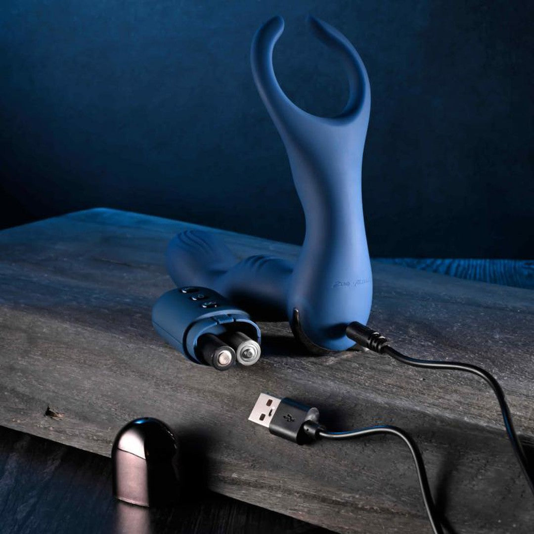 A Zero Tolerance By All Means Remote Control Prostate Vibrator, featuring a blue silicone design with a come-hither motion, is displayed on a wooden surface. It is accompanied by a black USB charging cable and an inconspicuous remote control. The dimly lit scene, set against a dark blue background, enhances the sense of mystery.