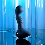 A sleek, dark-hued, curvy object with a smooth texture stands prominently on a reflective surface against a bright blue background. The Zero Tolerance Blue Oasis, a USB rechargeable vibrating silicone plug, is complemented by tall, clear glass cylinders that enhance the abstract and modern aesthetic of the scene.