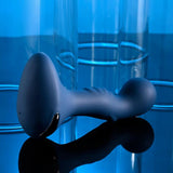 The Zero Tolerance Blue Oasis, a blue ergonomically shaped vibrating silicone plug with a curved design and glossy black accent, rests against a reflective surface. Surrounded by large, transparent blue tubes, the scene glows in cool ambient light. Experience 10 vibrating speeds with this USB rechargeable marvel from Zero Tolerance.