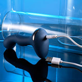 The Zero Tolerance Blue Oasis, an elegantly curved blue electronic device featuring USB rechargeability and 10 vibrating speeds, is connected to a USB charging cable. It is positioned on a reflective surface next to a transparent cylindrical object, all set against a tranquil blue backdrop.