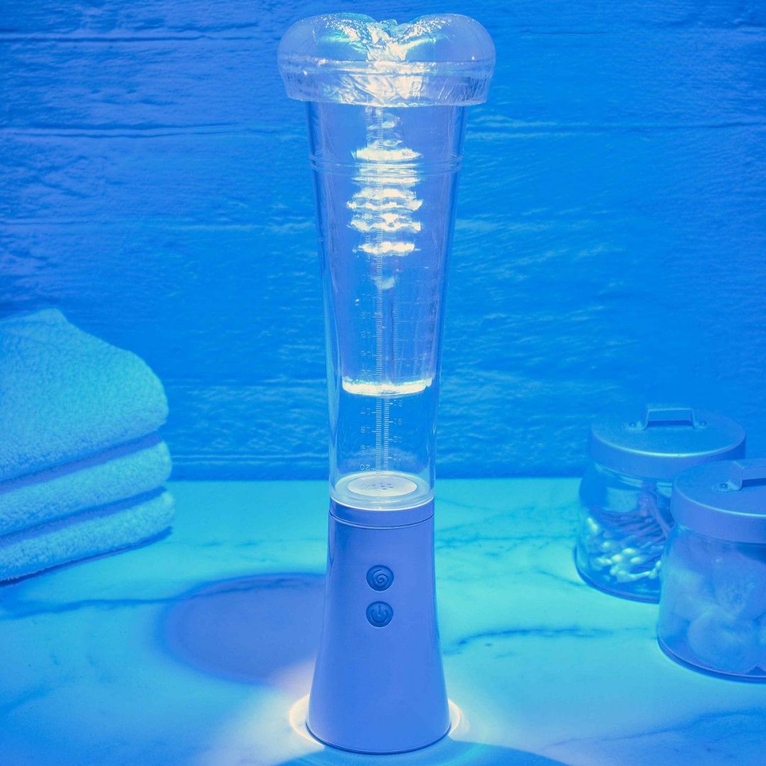 The Zero Tolerance Sucking Good Vibrating Masturbator, featuring a tall cylindrical design with a transparent upper section and controls on the white base, is filled with water and illuminated by a blue light. It rests on a marble surface near folded towels and jars, creating a serene ambiance.