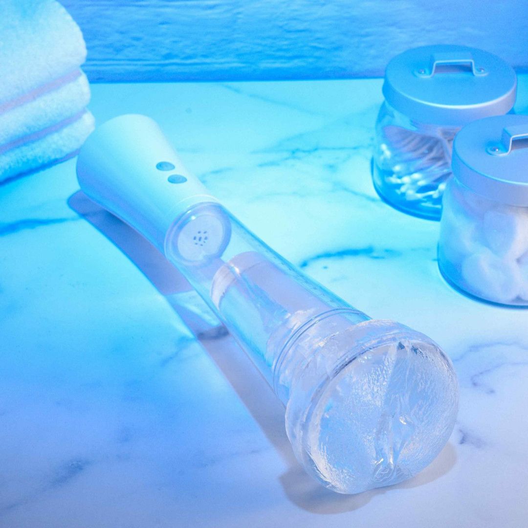 The image showcases a modern white showerhead with a transparent handle, elegantly placed on a marble surface. Nearby, neatly folded towels accompany two sleek glass jars containing cotton swabs and balls. The scene is enhanced by a calming blue light, reflecting the soothing experience offered by Zero Tolerance products like the Sucking Good Vibrating Masturbator.