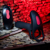 The Zero Tolerance Vibrating Girth Enhancer for Men, a black silicone ring-shaped device designed for vibrating penis enhancement, is showcased against a brick-patterned background illuminated with red lighting. Nearby lies a separate black oval remote, perfect for couple's play, both equipped with matching red power buttons.