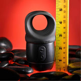 Beside a yellow measuring tape, suggesting exact measurements, the sleek black Zero Tolerance Wet it Be Lube Dispenser Stroker with its rounded loop on top stands prominently. Set against a red backdrop adorned with smooth black stones, this scene sparks curiosity, likely accentuated by the product's enticing multi-speed vibrations.