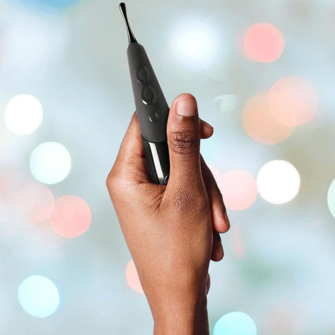 A hand holds the Zumio E Clitoral Stimulator by ZUMIO, INC—sleek, black, with control buttons—designed for precise stimulation and shown against a blurred backdrop of pastel lights.