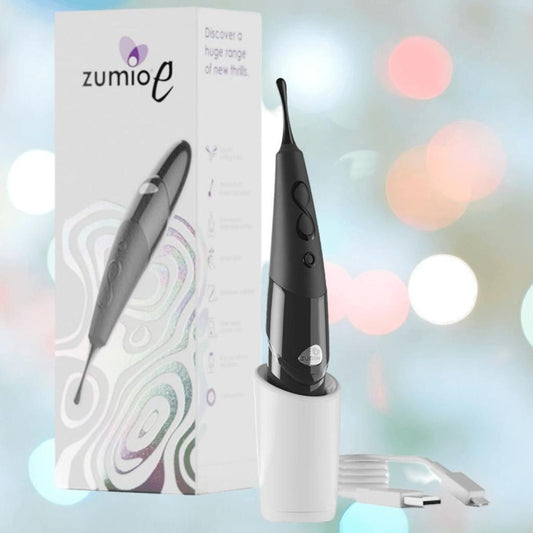 The Zumio E Clitoral Stimulator - Black by ZUMIO, INC stands upright in its white charging cup next to the product box, with a USB cable nearby. Soft pastel bokeh highlights Zumio E’s pressure-sensitive, precise stimulation features. 1080