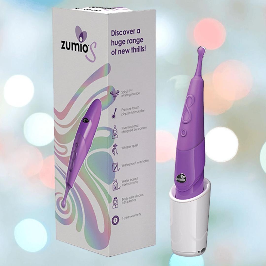 A light purple Zumio S Spirotip Clitoral Stimulator by ZUMIO, INC stands upright in its white charging base beside packaging with product info and colorful swirls on a soft, blurred background.