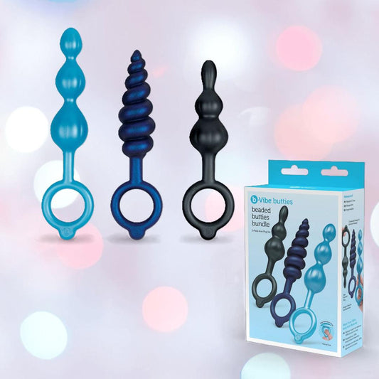 Three blue and black silicone butt plugs with finger loop handles from the b-Vibe Butties Bundle: Premium Butt Plug Kit with 3 Sizes are displayed upright by the packaging, highlighting this playful B-Vibe set against a soft pastel bokeh background. 1080