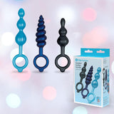 Three blue and black silicone butt plugs with finger loop handles from the b-Vibe Butties Bundle: Premium Butt Plug Kit with 3 Sizes are displayed upright by the packaging, highlighting this playful B-Vibe set against a soft pastel bokeh background.
