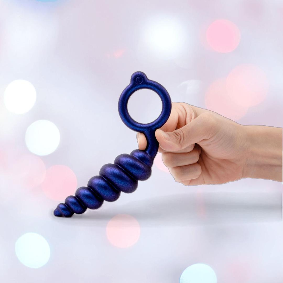A hand holds a dark blue silicone butt plug with a looped handle from the B-Vibe Butties Bundle: Premium Butt Plug Kit with 3 Sizes by B-Vibe, set against a soft, blurred background featuring pink, white, and blue bokeh lights.