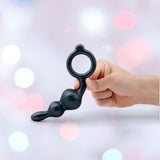 A hand holds a black silicone butt plug with a ring handle and three rounded beads—ideal for fans of the B-Vibe Butties Bundle: Premium Butt Plug Kit with 3 Sizes from B-Vibe, shown against a soft, pastel-lit background.