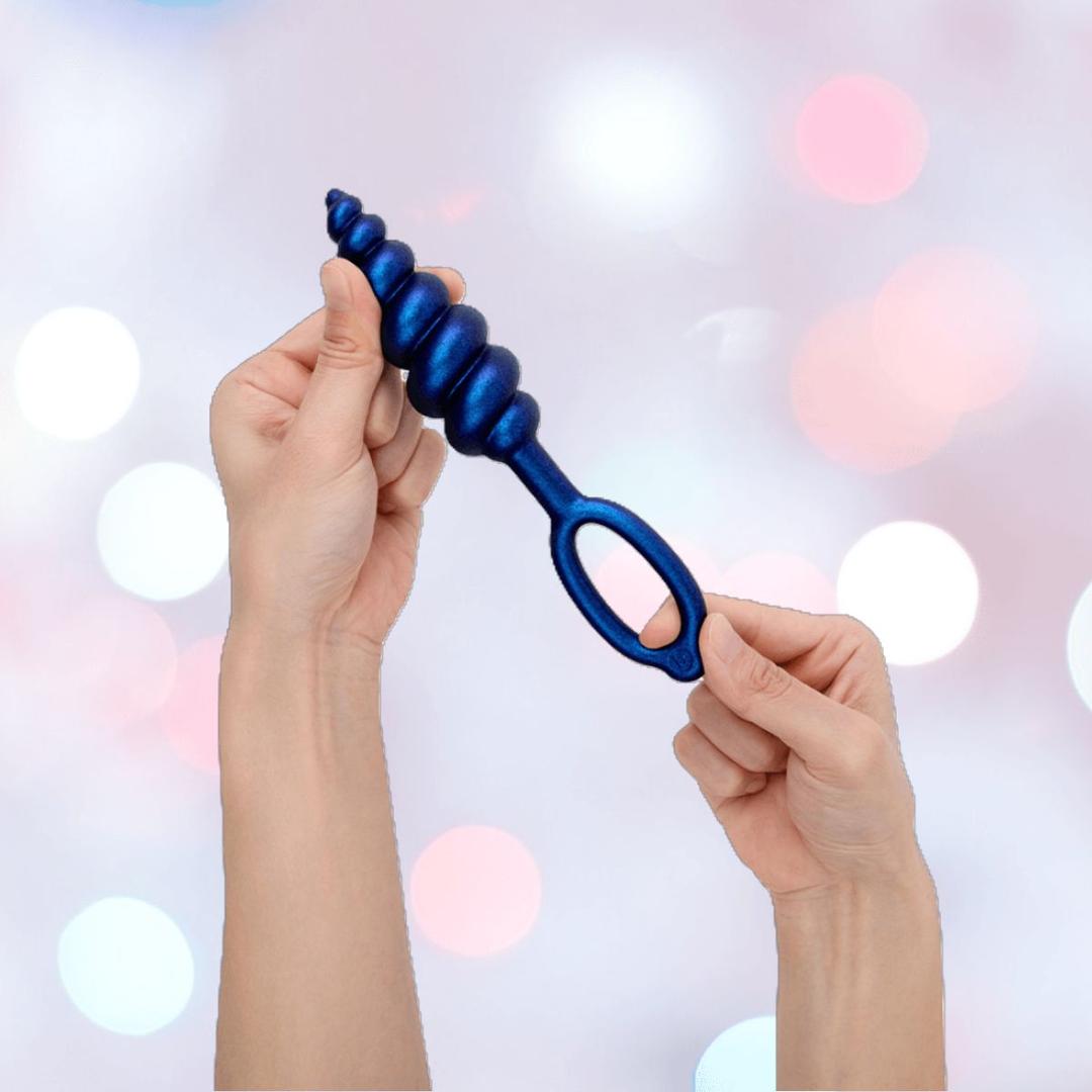 Two hands hold a blue b-Vibe Butties butt plug from the B-Vibe Butties Bundle: Premium Butt Plug Kit with 3 Sizes, featuring a ribbed silicone design and handle, against a soft pastel background.