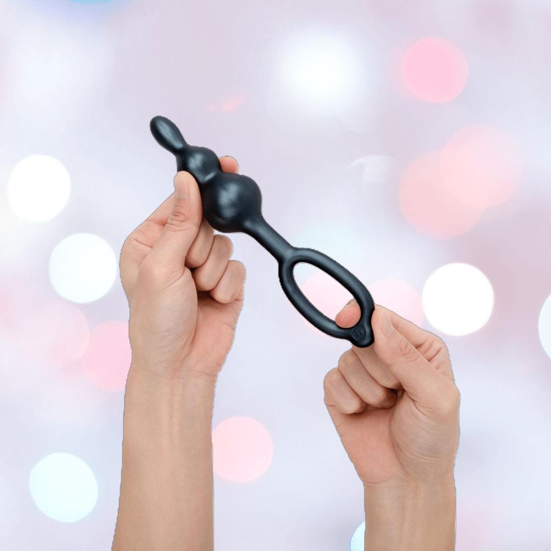 Two hands hold the B-Vibe Butties Bundle: Premium Butt Plug Kit with 3 Sizes, each with rounded bulbs and looped handles, against a pastel background with white and pink bokeh lights.