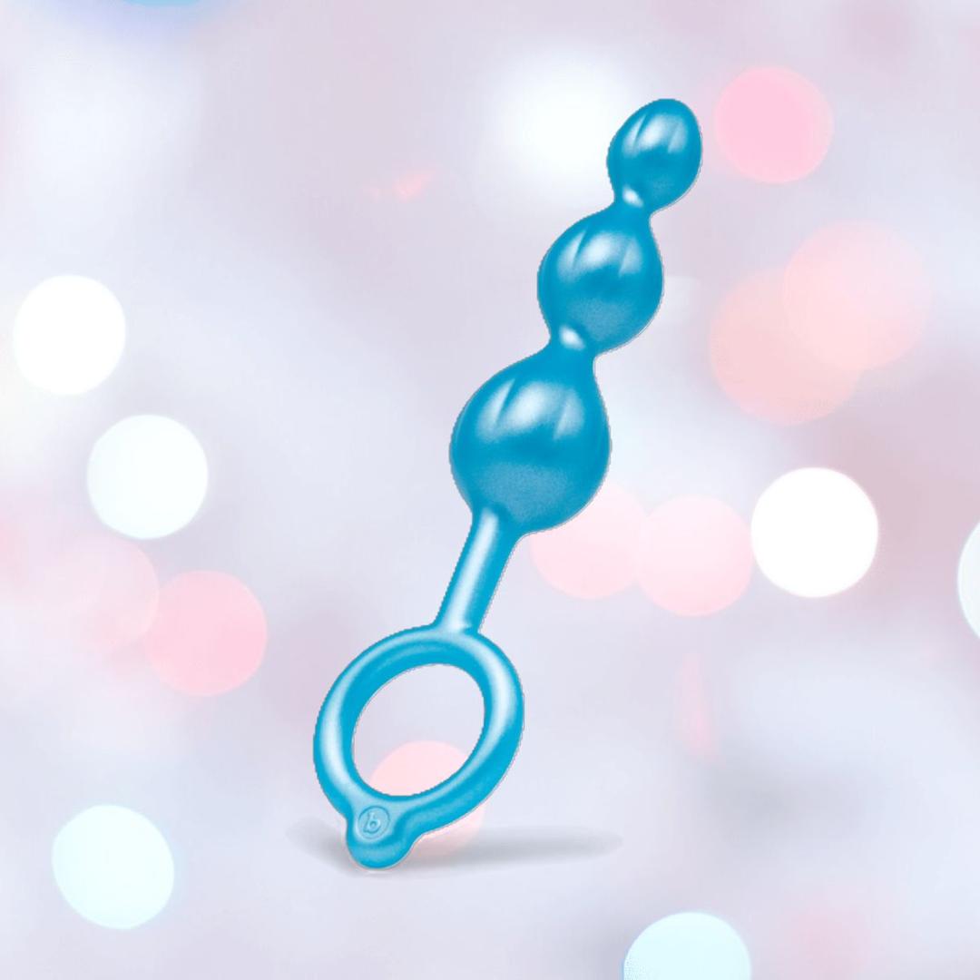 A B-Vibe Butties Bundle: Premium Butt Plug Kit with 3 Sizes features three graduated silicone plugs and a looped handle, shown against a soft pastel bokeh background.