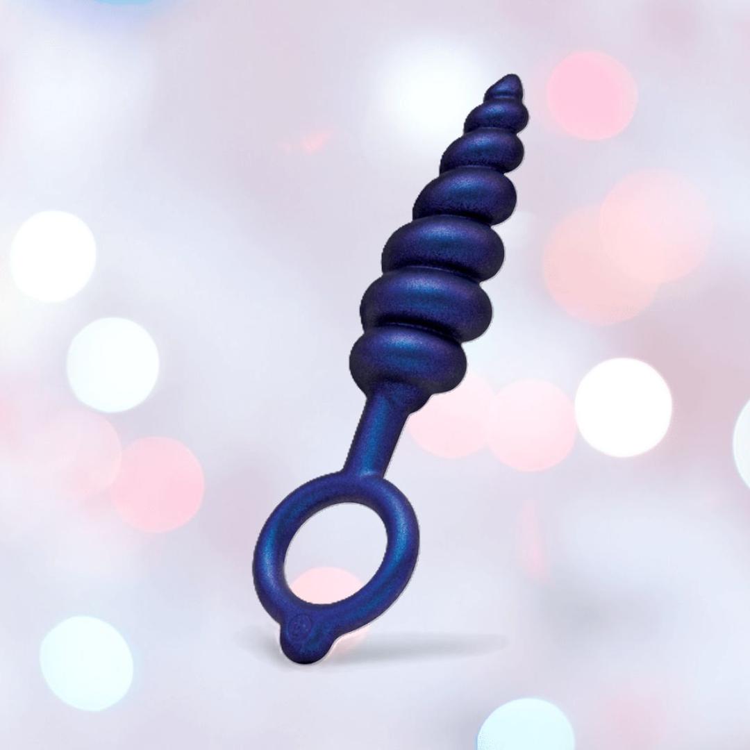 The b-Vibe Butties Bundle by B-Vibe is a premium silicone butt plug kit featuring 3 spiral-shaped, dark blue plugs with rounded tips and ring handles, shown against a soft background with white and pink bokeh lights.