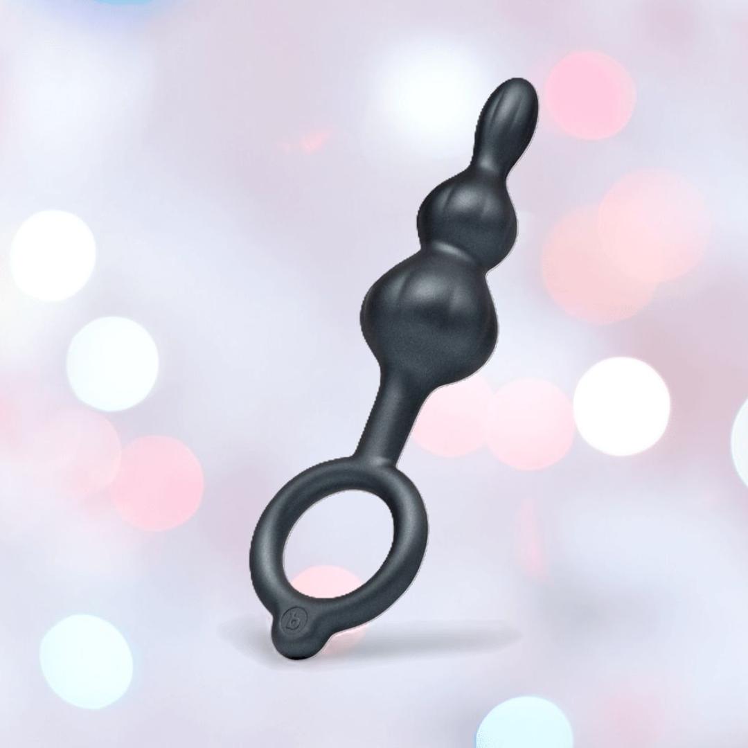 The b-Vibe Butties Bundle: Premium Butt Plug Kit with 3 Sizes by B-Vibe features black silicone plugs with graduated bulbs and a circular handle, shown against a pastel bokeh background of pink, blue, and white light spots.