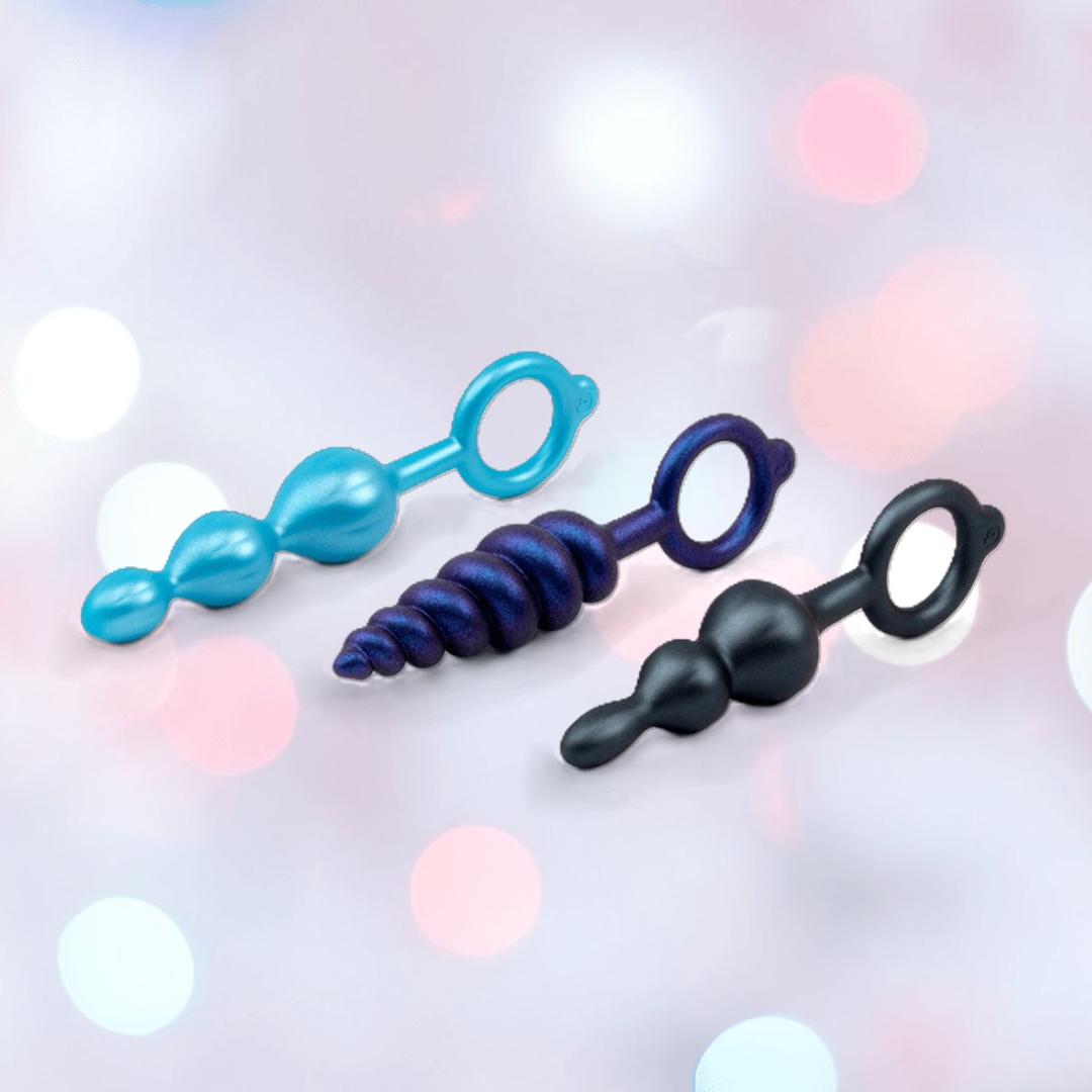 The b-Vibe Butties Bundle by B-Vibe includes three premium silicone butt plugs in blue and black, with ring handles—beaded, spiral, and dual-bulb styles—displayed on a soft pink and white background.