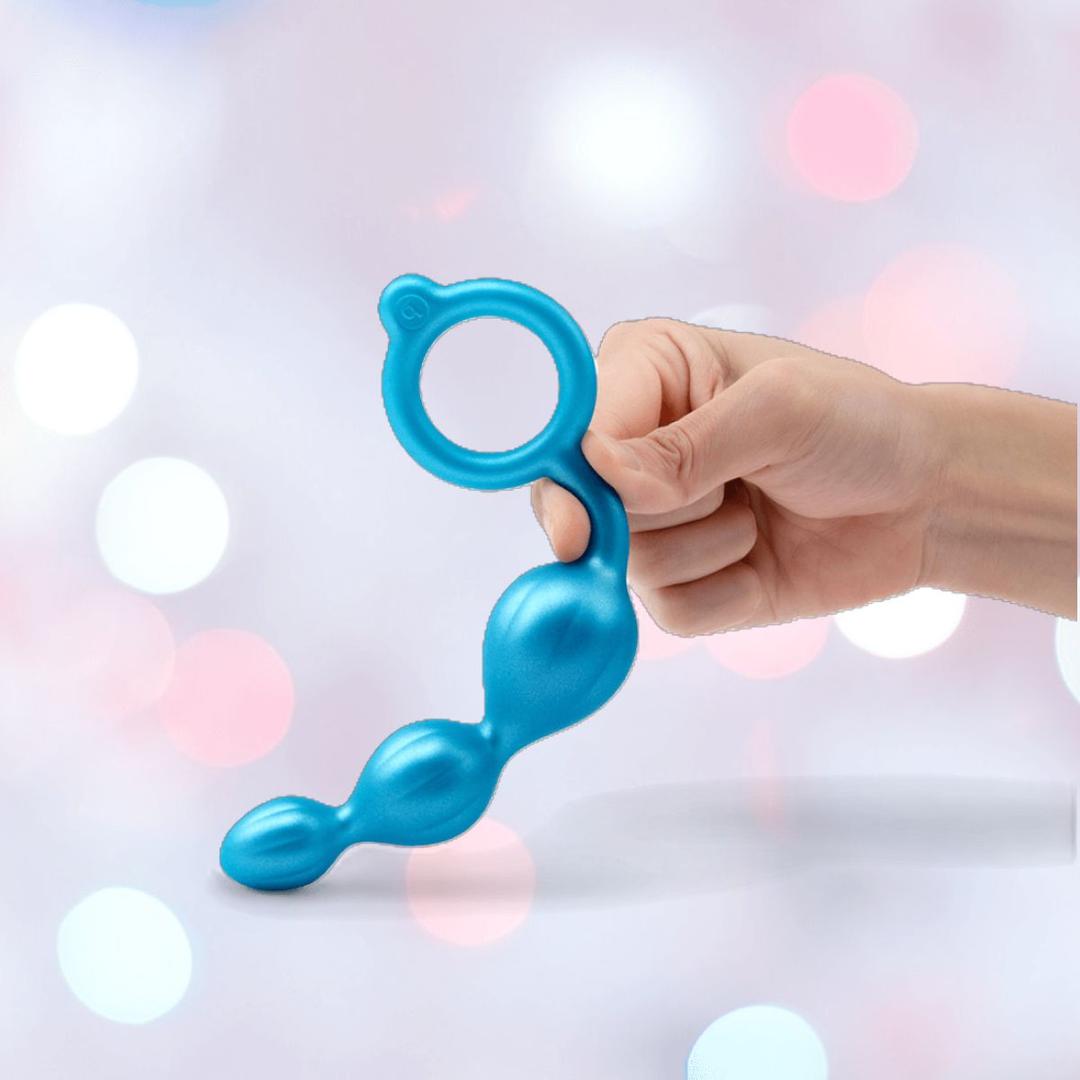 A hand holds a blue B-Vibe Butties Bundle anal plug with three bulbs and a looped handle, set against soft pink, white, and blue bokeh lights—an ideal choice from B-Vibe’s premium butt plug kit with 3 sizes.