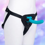 A close-up image features a person wearing the B-Vibe Every Body Strap-On Harness Set with a 6" silicone dildo by B-Vibe, set against a softly blurred pastel background.