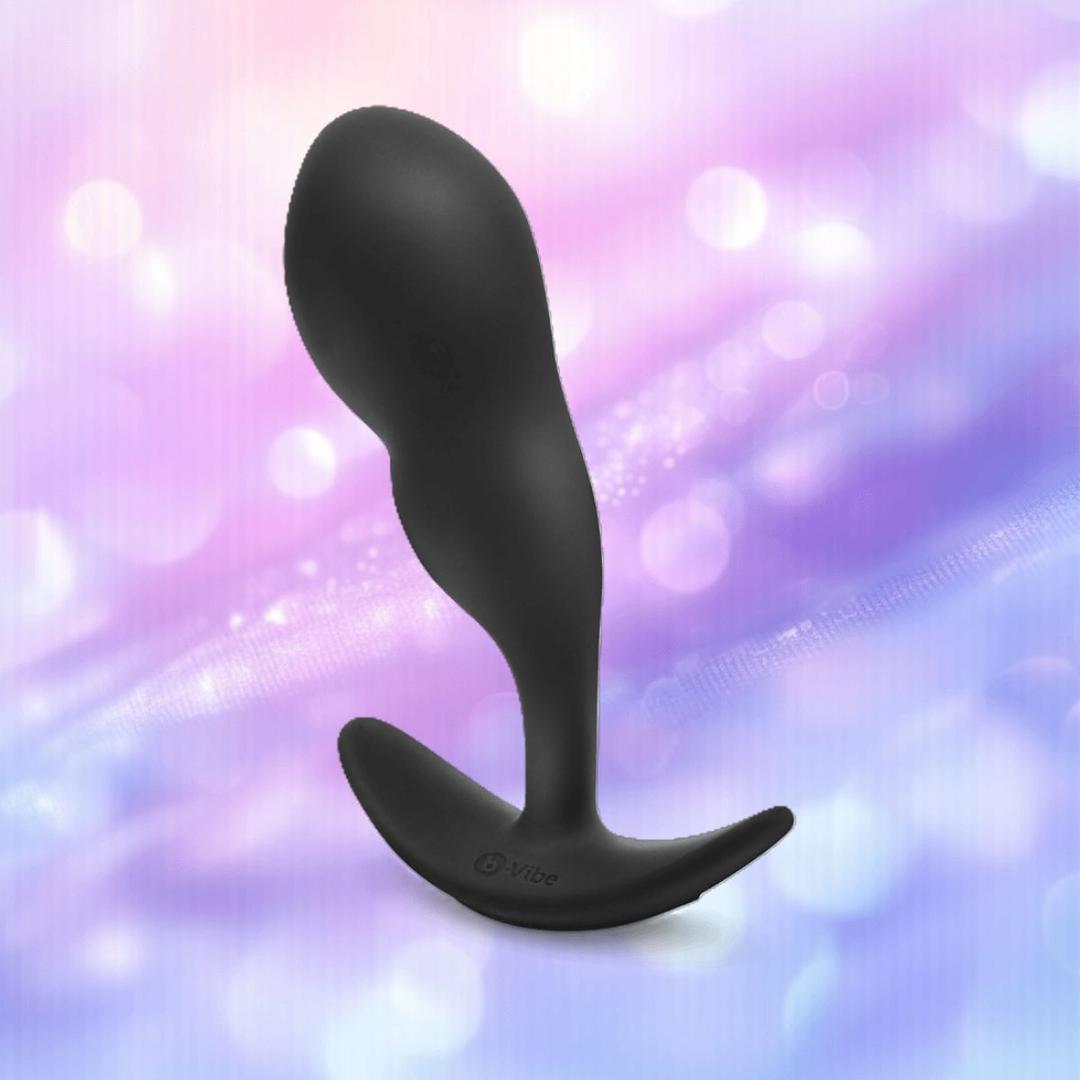 The B-Vibe Vibrating P-Spot Snug Plug RC Large by B-Vibe, in sleek black silicone and curved design, stands upright against a softly blurred pastel pink and purple background with glowing bokeh lights.
