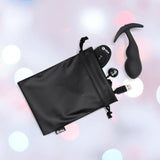 A black drawstring pouch holds a USB cable, remote control, and the B-Vibe Vibrating Snug Plug RC: Silicone P-Spot Anal Vibrator - Large by B-Vibe, all displayed on a blurred pastel background.