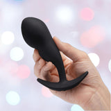 A hand holds the B-Vibe Vibrating Snug Plug RC: Silicone P-Spot Anal Vibrator - Large by B-Vibe, a black silicone plug with a flared base, set against a softly blurred pastel background.