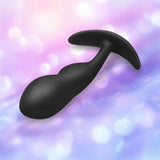 A B-Vibe Vibrating P-Spot Snug Plug RC Large, black silicone butt plug with a curved, ergonomic shape and flared base, is shown against a soft, blurred pastel background featuring hues of pink, purple, and blue.
