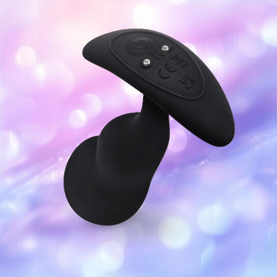The B-Vibe Vibrating P-Spot Snug Plug RC Large, a black, curved plug with control buttons from B-Vibe, is displayed against a soft pastel background with bokeh light effects.
