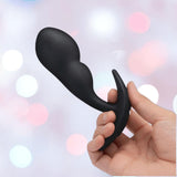 A hand holds the B-Vibe Vibrating Snug Plug RC, a large black silicone P-Spot anal vibrator by B-Vibe, against a softly blurred pastel bokeh background.