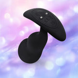 The B-Vibe Vibrating P-Spot Snug Plug RC Large, a black, curved plug with control buttons from B-Vibe, is displayed against a soft pastel background with bokeh light effects.