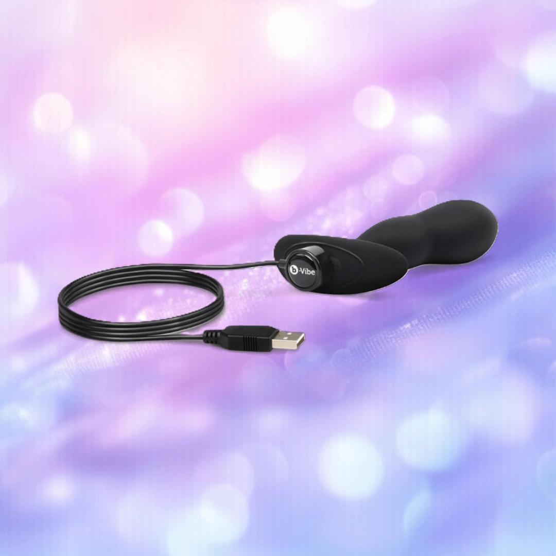 The B-Vibe Vibrating P-Spot Snug Plug RC Large by B-Vibe, a black curved USB-powered plug with a long cord, is displayed against a soft pastel purple and pink blurred background.