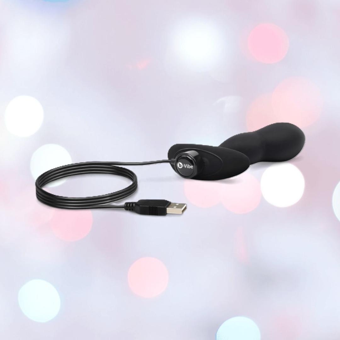 The B-Vibe Vibrating Snug Plug RC: Silicone P-Spot Anal Vibrator - Large by B-Vibe, shown in black with a USB charging cable attached, is displayed against a soft pastel background with blurred circular lights.