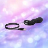 The B-Vibe Vibrating P-Spot Snug Plug RC Large by B-Vibe, a black curved USB-powered plug with a long cord, is displayed against a soft pastel purple and pink blurred background.